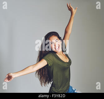 Vivacious attractive casual young woman celebrating throwing her arms in the air and laughing at the camera, over grey in square