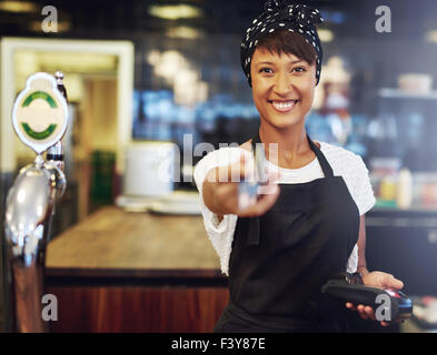 Vivacious attractive young African American bar tender handing back a bank card after processing payment conceptual of a small b Stock Photo
