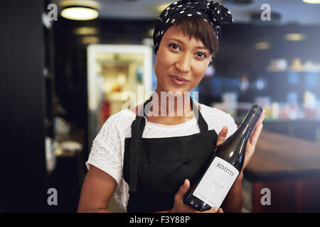 Attractive young African American lady in an apron presenting a bottle of red wine to a customer in a restaurant or bar before o Stock Photo