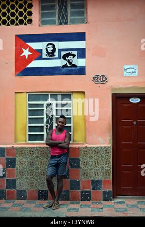 portrait of a young Cuban man standing with folded arms outside colorful home with pink walls, faded tiles and Cuban flag mural Stock Photo