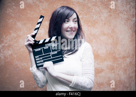 Young woman in 70s hippie style smiling with clapperboard, outdoor orange wall background Stock Photo