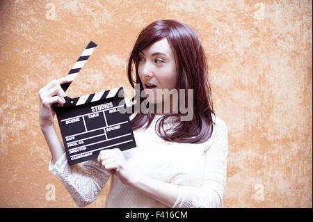 Young woman in 70s hippie style smiling with clapperboard, outdoor orange wall background Stock Photo