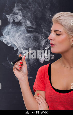 Stylish blond woman smoking an e-cigarette exhaling a cloud of smoke with her eyes closed in enjoyment, profile view on a dark b Stock Photo