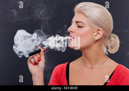 Stylish blond woman smoking an e-cigarette exhaling a cloud of smoke with her eyes closed in enjoyment, profile view on a dark background Stock Photo