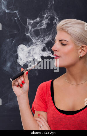 Stylish blond woman smoking an e-cigarette exhaling a cloud of smoke with her eyes closed in enjoyment, profile view on a dark background Stock Photo