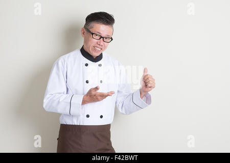 Portrait of 50s mature Asian male chef in uniform showing something and giving thumb up, standing on plain background with shado Stock Photo