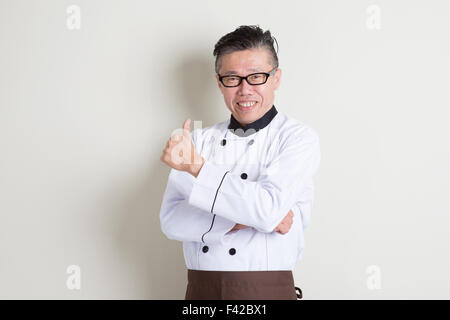 Portrait of confident 50s mature Asian male chef in uniform arms crossed and thumb up, standing on plain background with shadow, Stock Photo