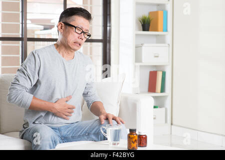 Mature 50s Asian man stomachache,  pressing on stomach with painful expression, sitting on sofa at home, medicines on table. Stock Photo