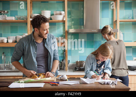 Young daughter drawing ar kitchen counter while parents prepare meal Stock Photo