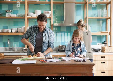 Young daughter drawing ar kitchen counter while parents prepare meal Stock Photo