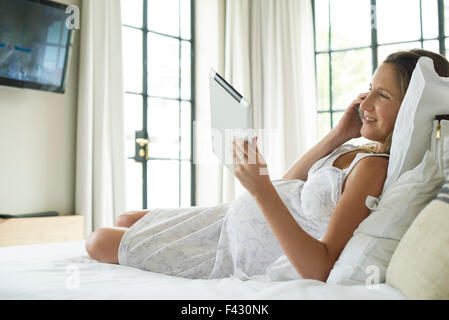 Pregnant woman relaxing in bed, using digital tablet and chatting on cell phone Stock Photo