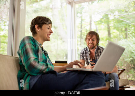 A man and woman sitting together in a room with large picture windows, one using a laptop. Stock Photo