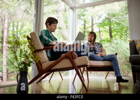 A man and woman sitting together in a room with large picture windows, one using a laptop. Stock Photo
