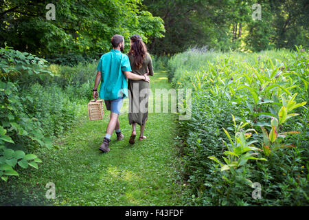 A man and woman walking through a meadow, through the long grass, carrying a picnic basket. Stock Photo