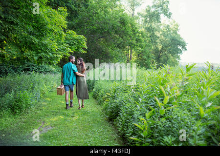 A man and woman walking through a meadow, through the long grass, carrying a picnic basket. Stock Photo