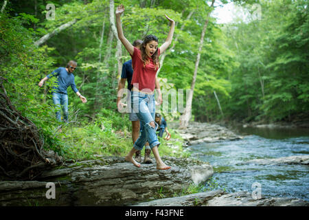 Two men and two women on the riverbank, one leaping across stepping stones. Stock Photo