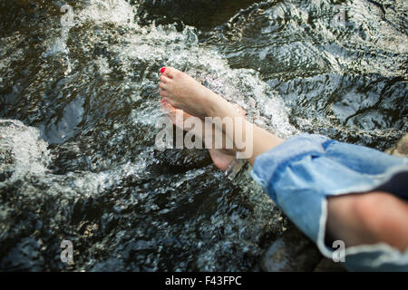 A woman in fashionable jeans with a rip, with her feet in the cool flowing waters of a river. Stock Photo