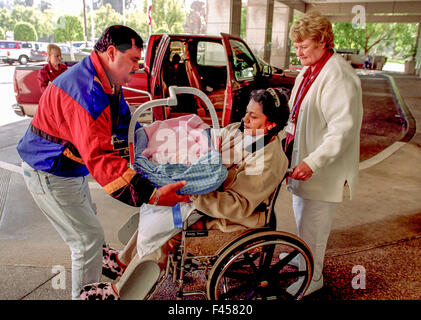 The Hispanic father of a newborn  carefully lifts a carry crib from his wife in a wheelchair outside a hospital in Mission Viejo, CA. Note volunteer at right and truck in background. Stock Photo