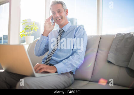 Businessman sitting on a couch having phone call and using laptop Stock Photo