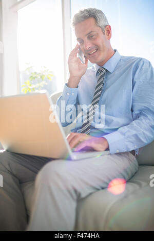 Businessman sitting on a couch having phone call and using laptop Stock Photo