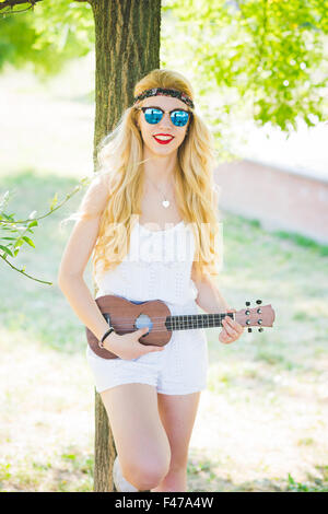 Knee figure of young handsome caucasian long blonde straight hair hippy woman posing leaning against a tree in a city park, playing a ukulele, looking in camera, smiling - music, inspiration concept Stock Photo