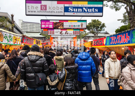 Ikuta Shinto shrine, Japan during Shogatsu, new year. Crowds walking along avenue to the shrine with food stalls on either side and over head adverts. Stock Photo