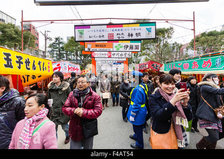 Ikuta Shinto shrine, Japan during Shogatsu, new year. Crowds walking along avenue to the shrine with food stalls on either side and over head adverts. Stock Photo