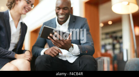 Happy young business people sitting together using digital tablet while at hotel lobby. Focus on tablet computer. Stock Photo
