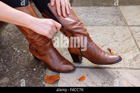 Woman zips up high-heeled tan leather boots, autumn leaves scattered at her feet