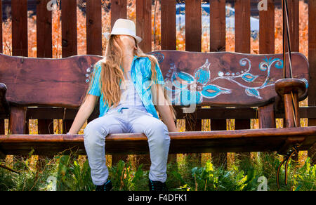 Blonde, 11 years old girl in blue shirt sitting on a garden swing, covering her face with a white hat. Autumn colors Stock Photo