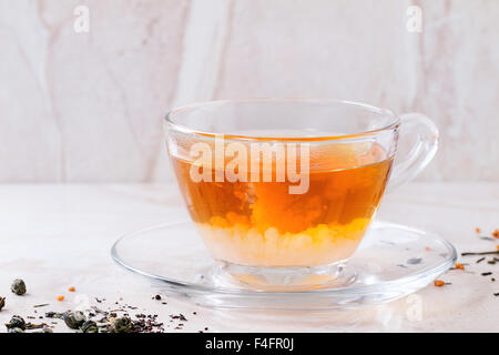 Milk dissolves in glass cup of hot tea on saucer with dry green and black tea leaves over white marble backgtound. Stock Photo