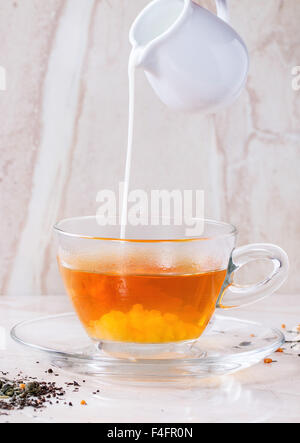 Milk pouring from white jug to glass cup of hot tea on saucer with dry green and black tea leaves over white marble backgtound. Stock Photo