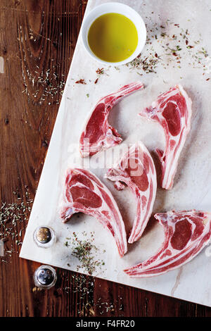 Raw lamb chops with salt, pepper, dry herbs and bowl of olive oil on marble board over wooden table. Top view. Stock Photo