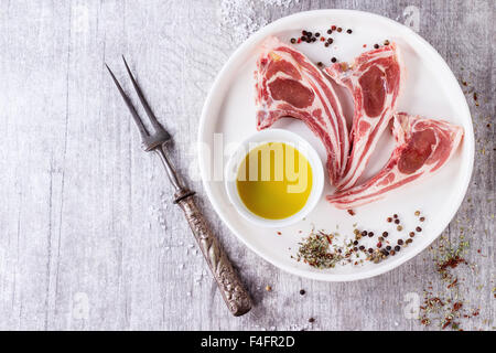 Raw lamb chops with salt, pepper, dry herbs and bowl of olive oil on white ceramic plate over white wooden table. Meat fork near Stock Photo