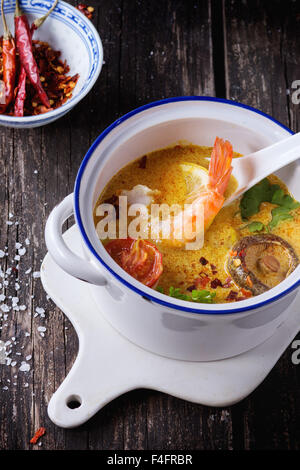 White Ceramic pan with Spicy Thai soup Tom Yam with Coconut milk, Chili pepper and Seafood on ceramic cutting board over old woo Stock Photo