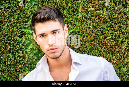 Handsome young man in elegant white shirt standing outdoor in front of green bushy hedge looking at camera Stock Photo