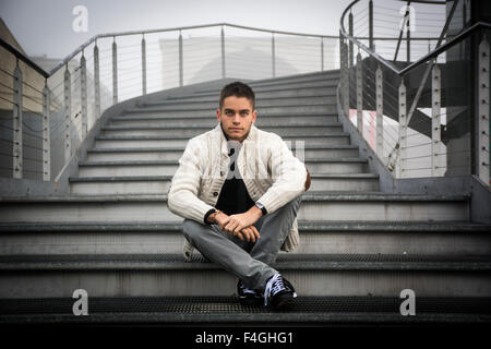 Well dressed handsome young man in casual clothes sitting cross legged on the lower part of a stairway outdoor in winter Stock Photo