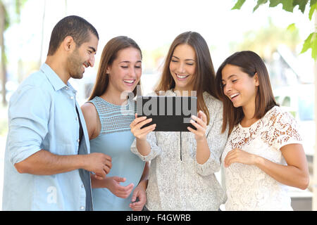 Front view of a group of four happy friends watching videos on a tablet in the street Stock Photo