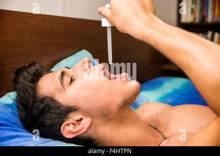 Sick or unwell young man using dropper pipette on his mouth for medicine, drug treatment, while lying in bed Stock Photo