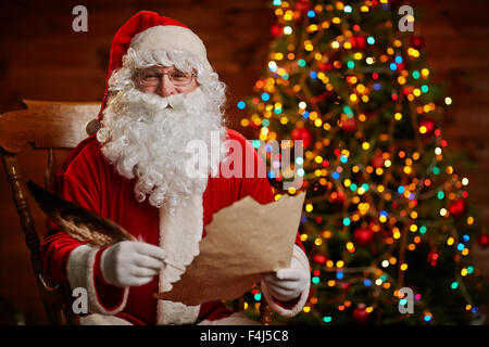 Cheerful Santa with paper and feather looking at camera while writing Christmas letter Stock Photo