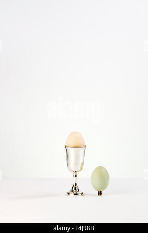 Two free range hens eggs, one green, one pale brown, one in a silver egg cup and the other precariously sitting on a tiny cup on a white background. Stock Photo