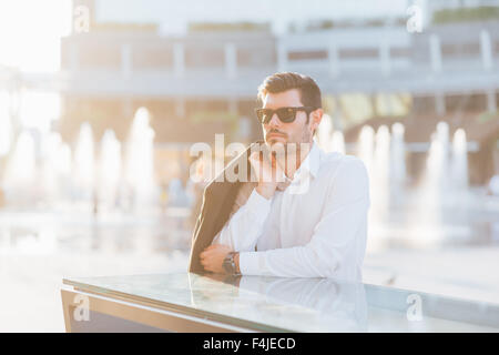 young handsome black hair modern businessman leaning on a counter in the city backlight, overlooking right, carrying his coat, wearing sunglasses - successful, business concept Stock Photo