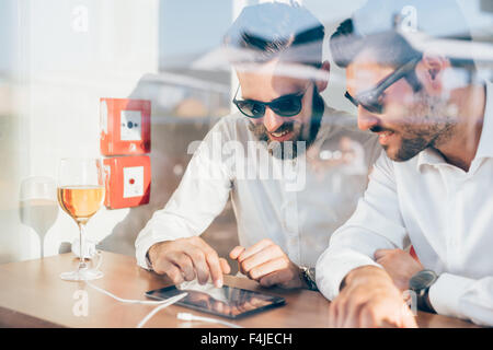 Two young bearded caucasian modern business man sitting in a bar, using tablet looking downward, touching the screen - business, work, technology concept - view behind a shop window Stock Photo