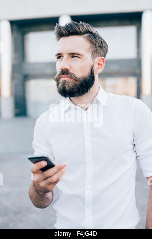 Half length of young handsome blonde caucasian modern businessman holding a smartphone, overlooking right, pensive - business, working, technology concept Stock Photo