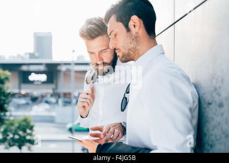 Half length of two young bearded blonde and black hair modern businessman, leaning against a wall, using tablet, looking downward the screen - technology, business, work concept Stock Photo