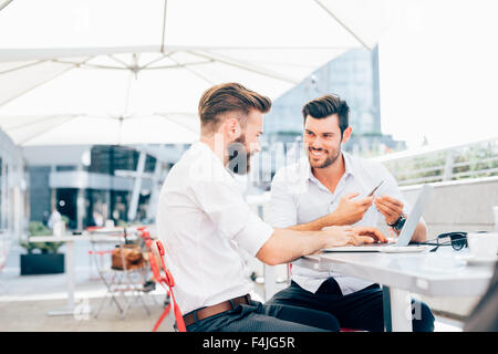 Two young bearded caucasian modern business man sitting in a bar, using smartphone and laptop, looking each other, smiling - business, work, technology concept Stock Photo