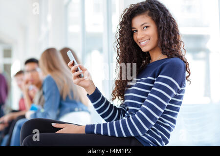 Portrait of a happy afro american female student using smartphone in university hall and looking at camera Stock Photo