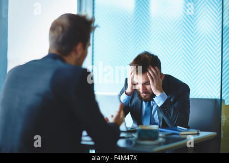 Male employee touching head while listening to his colleague at meeting Stock Photo