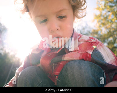 Landscape portrait of curly haired Caucasian toddler boy in a checked shirt outdoors with sun and trees in the background Stock Photo
