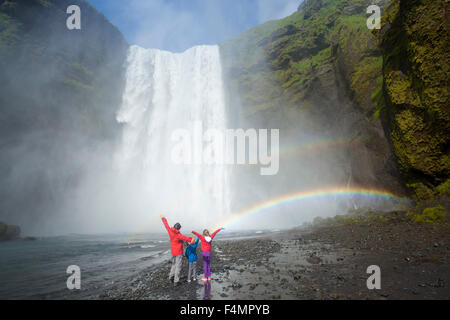 Family in the rainbow beneath 60m-high Skogafoss waterfall, Skogar, Sudhurland, Iceland. Stock Photo